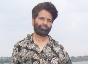 Portrait of Dr. Afroz Taj. Dr. Taj is an Indian man with straight black hair and a black beard. He is wearing a leaf-patterned shirt and standing in front of a waterfront.