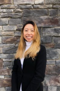 Portrait of Amy Lo. Amy is an Asian woman with long blond hair. She is wearing a black jacket over a white turtleneck and smiling in front of a gray brick wall.