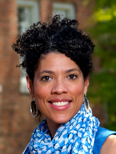 Portrait of Dr. Amy Locklear Hertel. Dr. Locklear Hertel is a multiracial woman with black curly hair. She is wearing large hoop earrings, a blue patterned scarf and a blue blouse and is standing outside smiling at the camera.