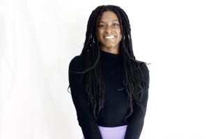 Portrait of Aneesha Tucker. Aneesha is a Black woman with long dreadlocks. She is wearing a black shirt, purple skirt, and gold hoop earrings and nose ring. She is smiling at the camera. 