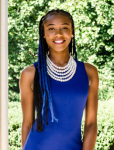 Portrait of Angum Check. Angum is a Black woman with blue and black braids. She is wearing a blue dress, a five-layer pearl necklace, and earrings and is standing outside smiling at the camera.