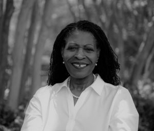 Portrait of Betsy Ayankoya. Betsy is a Black woman with straight black hair. She is wearing a white collared shirt and pearls and standing outside, smiling at the camera.