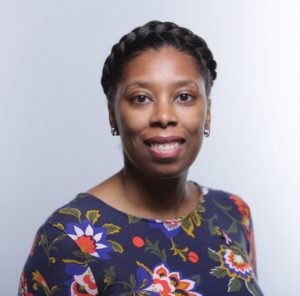 Portrait of Dawna Jones. Dawna is a Black woman with a halo of braids. She is wearing a floral patterned shirt, small silver hoop earrings and is smiling at the camera.