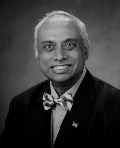 Portrait of Deb Aikat. Deb is a South Asian man with white hair. He is wearing a suit with a striped bow tie and American flag lapel pin and smiling at the camera.