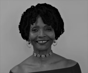 Portrait of Dr. Devetta Holman-Copeland. Dr. Holman-Copeland is a Black woman with short curly hair. She is wearing gold earrings, a metal choker, and a red off-the-shoulder blouse. She is smiling at the camera.