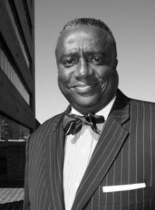 Portrait of Eric Watson. Eric is a Black man with short salt-and-pepper hair. He is wearing a pinstripe suit, white dress shirt and bowtie and is standing beside a skyscraper.
