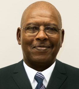 Portrait of Franklin Seymore. Franklin is a Black man with a bald head. He is wearing wire-rimmed glasses, a black suit, a white dress shirt and a blue striped tie.
