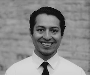 Portrait of Geovani Ramirez. Geovani is a latino man with short brown hair. He is wearing a white collared shirt and dark tie and smiling in front of a brick wall.
