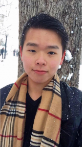 Portrait of Jacky Zheng. Jacky is an Asian man with short black hair. He is wearing a brown plaid scarf and black sweater and standing against a tree trunk surrounded by snow.