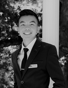 Portrait of Johnny Vang. Johnny is an asian man with gelled black hair. He is wearing a suit and tie and leaning against a pole, smiling.