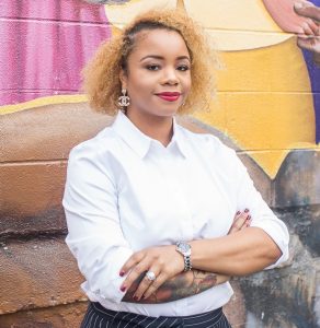Portrait of Dr. Khalilah R. Johnson. Dr. Johnson is a Black woman with short curly blond hair. She is wearing a white dress shirt and standing in front of a mural with her arms folded.