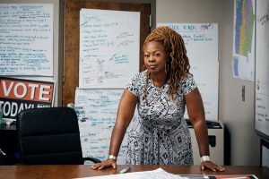Portrait of LaTosha Brown. LaTosha is a Black woman with long amber dreadlocks. She is wearing a black and white patterned dress, silver hoop earrings and silver bangle bracelets. She is standing in a conference room surrounded by sheets of paper with brainstorming notes. Her hands are on the table in an active stance and she is looking at the camera.