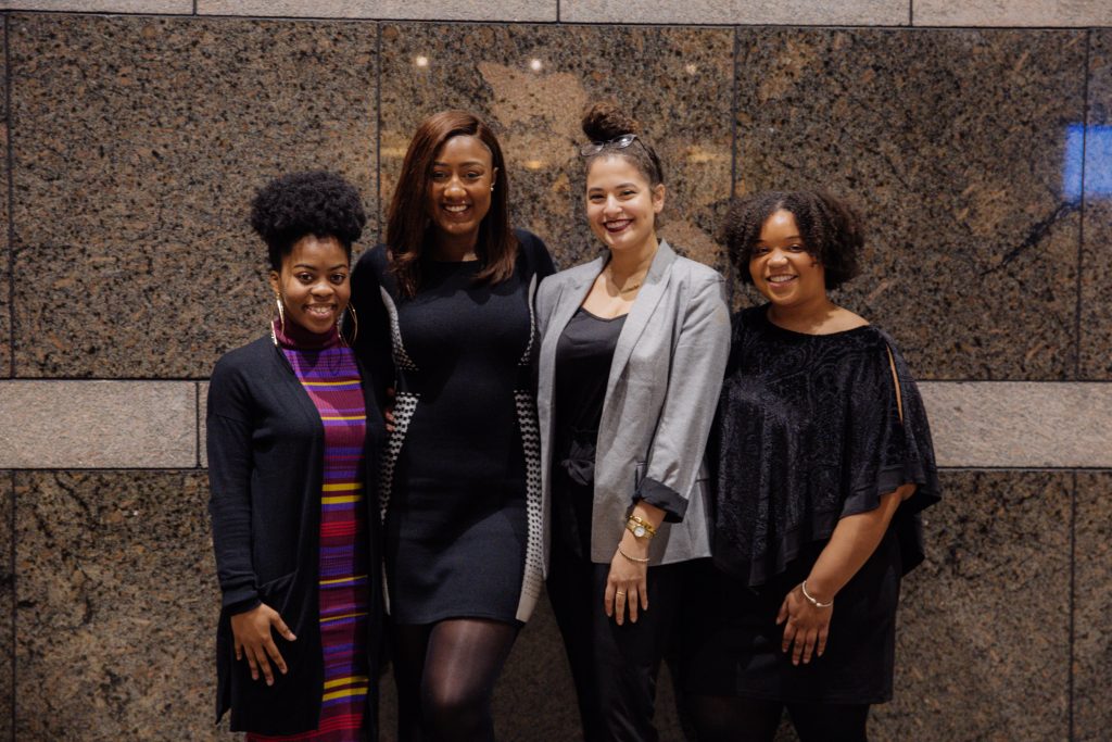 Photo of 2018-2019 MLK Committee members. There are four black women in the photo smiling at the camera and standing in front of a brown marble wall.