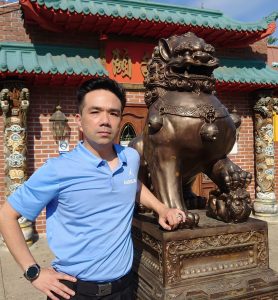 Portrait of Marcus Donie. Marcus is an asian-american man with short black hair. He is wearing a Carolina blue polo shirt and standing next to a lion statue.