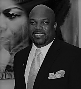 Portrait of O.J. McGhee. O.J. is a black man with a bald head and a black goatee. He is wearing a suit and standing in front of a poster.