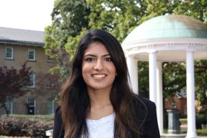 Portrait of Rhea Bhagia. Rhea is an Indian American woman with long straight brown hair. She is wearing a blue suit jacket and white shirt and smiling in front of the Old Well.