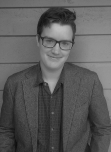 Portrait of Stephen Krueger. Stephan is a white man with short black hair. He is wearing glasses, a button down shirt, and a jacket and standing in front of a wall.