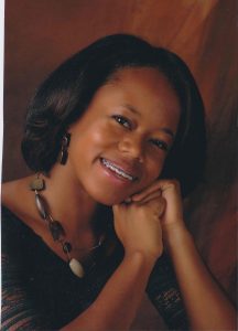 Portrait of Trinity Johnson. Trinity is a Black woman with a straight black bob. She is wearing a chunky necklace and earrings and a gray shirt and resting her head in her hands as she smiles at the camera.