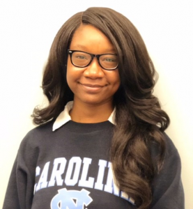 Portrait of Erica Wallace. Erica is a Black woman with long wavy hair. She is wearing a Carolina sweatshirt, white collared shirt and black-rimmed glasses and smiling at the camera.