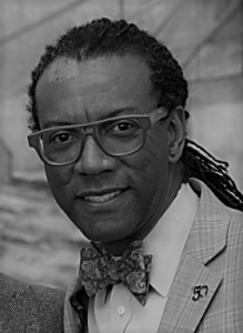 Portrait of Kenneth Ward. Kenneth is a black man with dreadlocks tied back in a ponytail. He is wearing glasses and a suit with a bowtie and smiling at the camera.