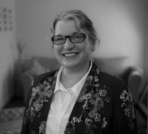 Portrait of Echo Meyer. Echo is a white woman with wavy gray hair. She is wearing glasses, a collared shirt and a floral suit jacket and smiling at the camera.